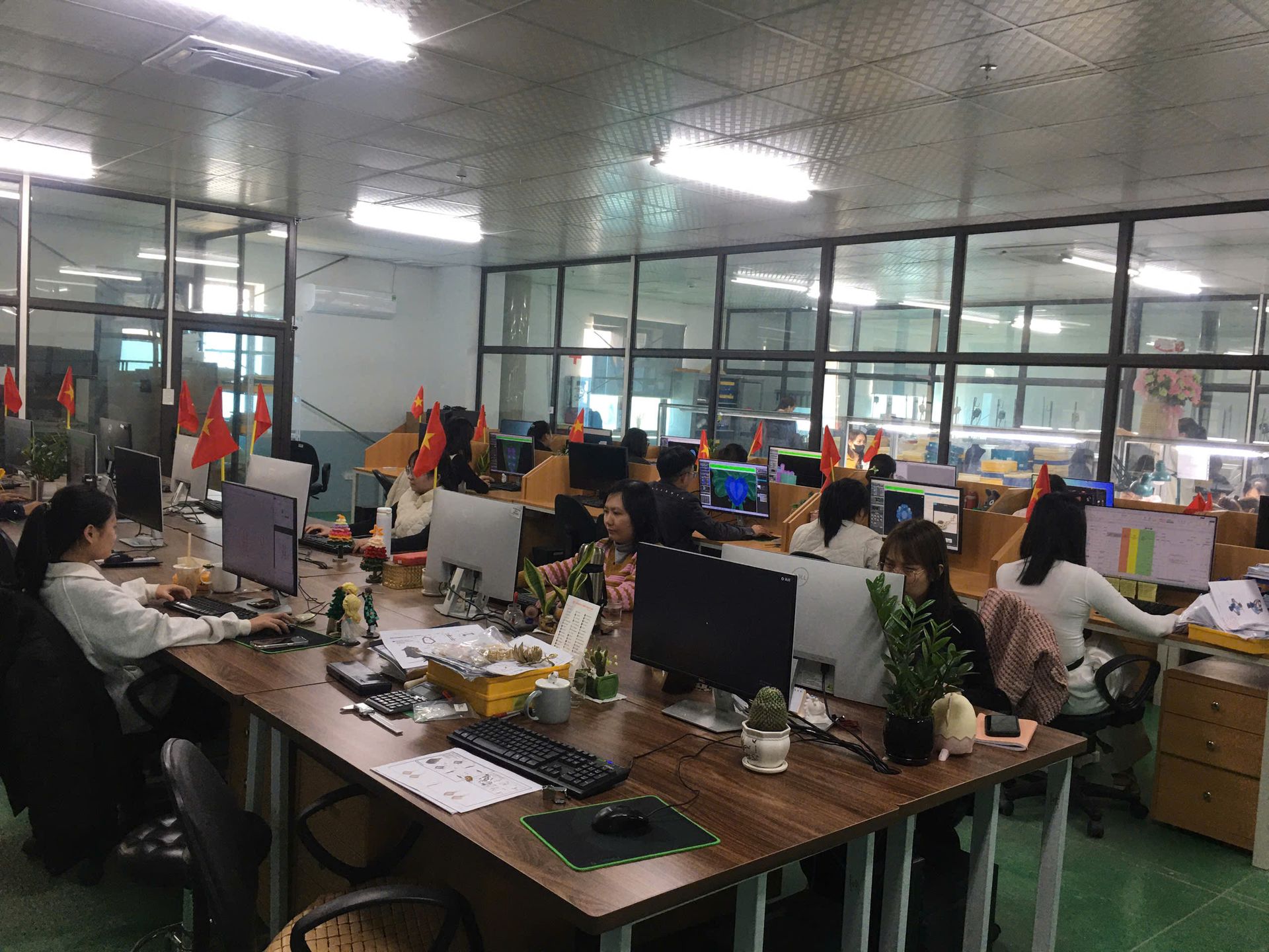 A wide-angle interior photograph of a bustling, collaborative open-plan office, as referenced in the file '1.jpg'. Multiple employees are seated at a long wooden communal desk, each working on individual desktop computers. The office environment is bright, illuminated by overhead fluorescent lights. Several small Vietnamese flags are displayed along the desk and cubicle dividers. The background features a large glass-partitioned area, offering a view into an adjacent, well-lit manufacturing or workshop space. The overall atmosphere is productive and professional.
